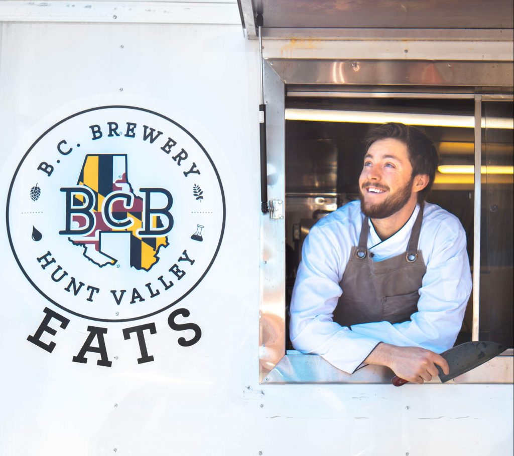 pic of Chef Dave in the window of BCB Eats food truck, the BCB sign to his right. he has a chef shirt and apron on and a chef's knife in his hand